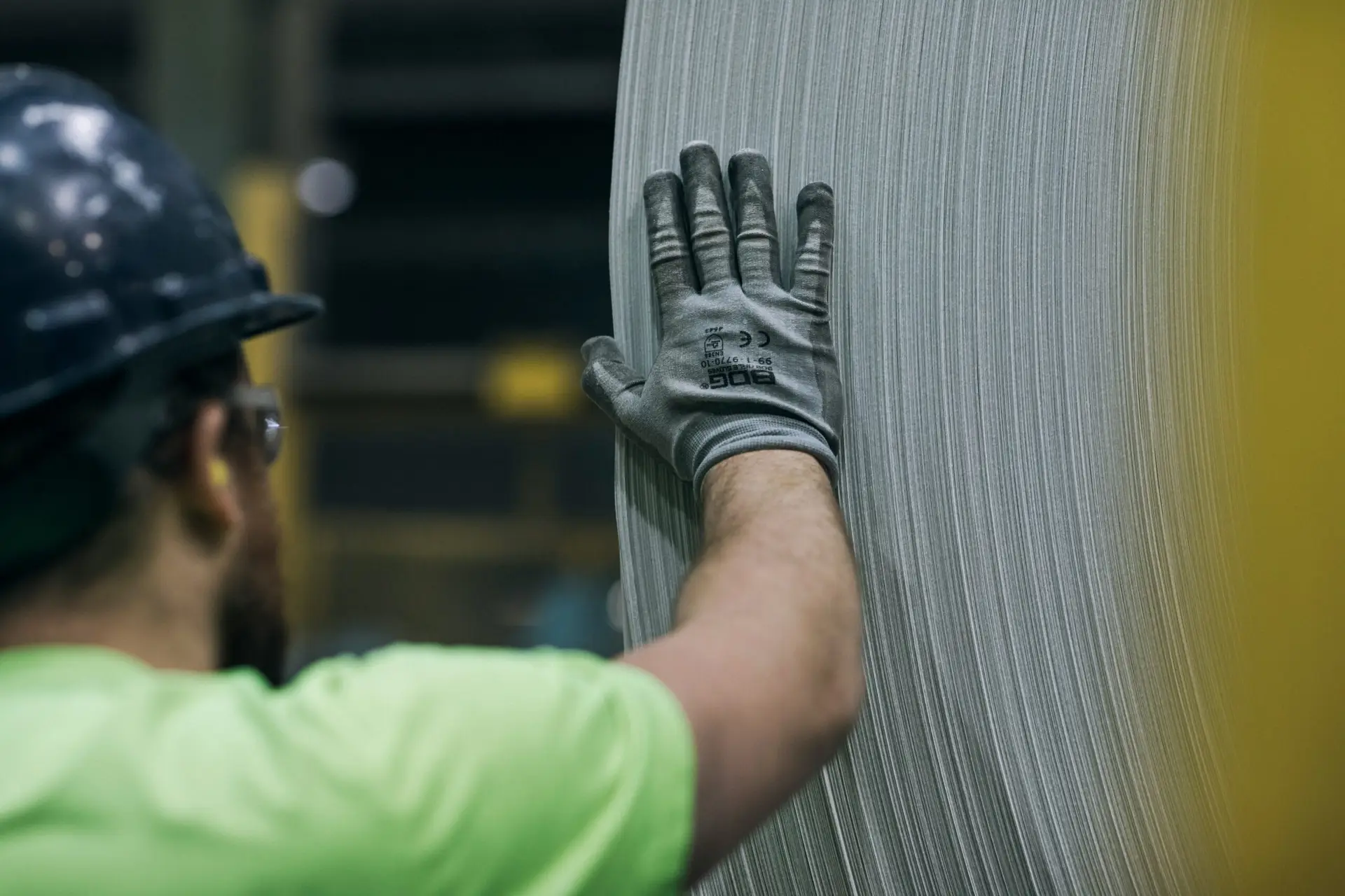 Factory worker wearing a helmet and gloves, inspecting a large machine component.